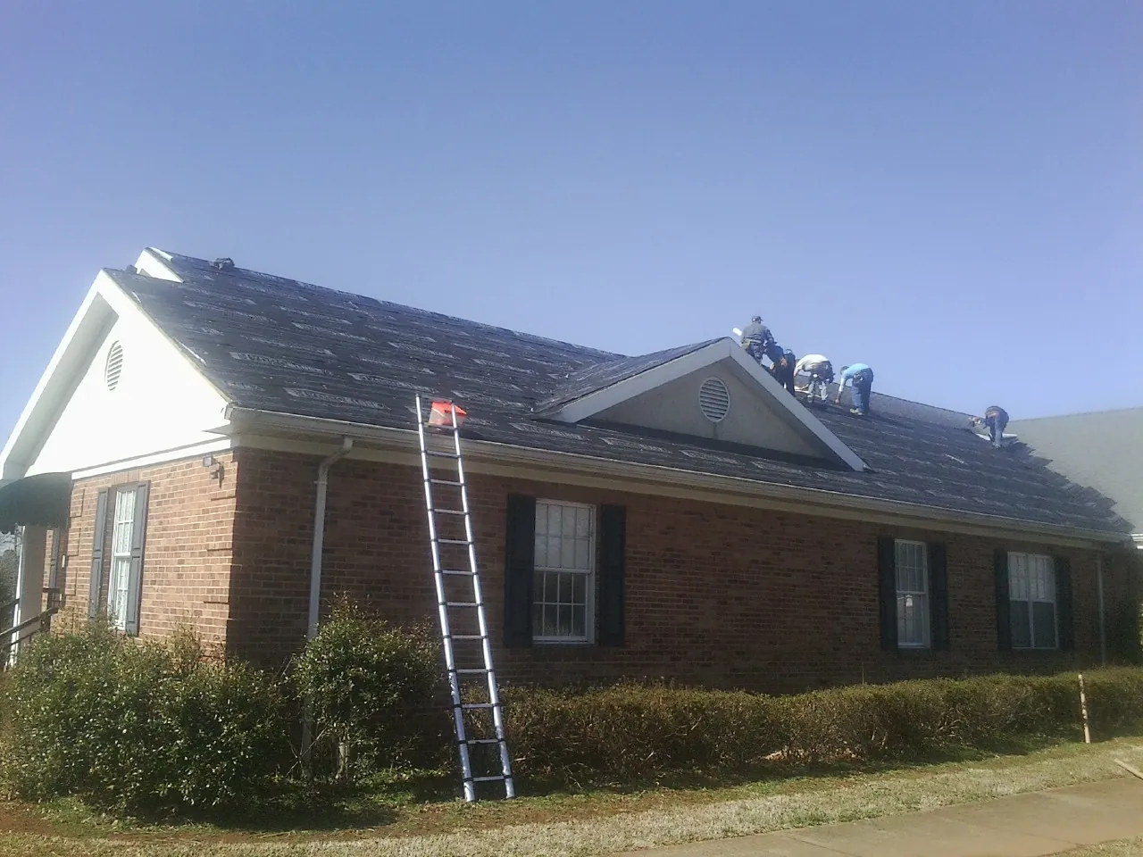 Crew tearing shingles off a brick ranch roof with a ladder set — Metal Roof Repair work by our crew in Redfield